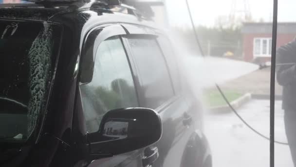 Mężczyzna myje czarny samochód. Slow Motion Video z procesu mycia samochodu na samoobsługowej myjni samochodowej. Jet wody z wysokim ciśnieniem zmyć brud z samochodu. Widok boku. Odpływy detergentów spienionych - Materiał filmowy, wideo