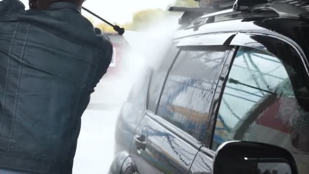 Ein Mann wäscht ein schwarzes Auto. Zeitlupenvideo einer Autowäsche an einer Selbstbedienungswaschanlage. Ein Wasserstrahl mit hohem Druck wäscht den Schmutz vom Auto ab. Seitenansicht. Abflüsse von geschäumtem Waschmittel - Filmmaterial, Video