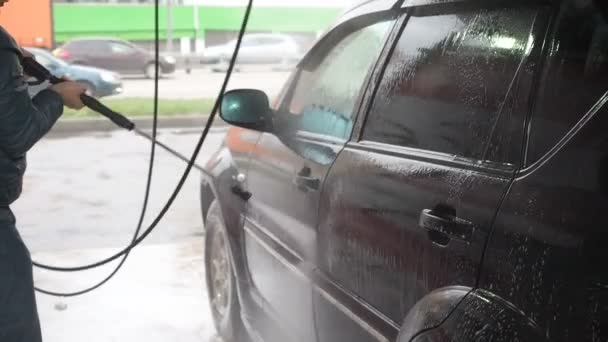 Muž myje černé auto. Zpomalený pohyb Video procesu mytí auta na samoobslužné mytí automobilu. Proud vody s vysokým tlakem smyjte ze špíny z auta. Side View. Odvodnění pěnového pracího prostředku - Záběry, video