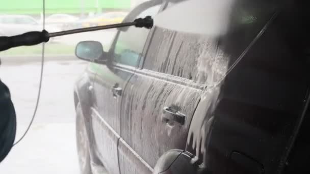 Ένας άντρας πλένει ένα μαύρο αυτοκίνητο. Αργή κίνηση βίντεο από μια διαδικασία πλύσης αυτοκινήτων σε ένα αυτο-service πλυντήριο αυτοκινήτων.Μια αεριωθούμενη του νερού με ένα πλύσιμο υψηλής πίεσης από τη βρωμιά από το αυτοκίνητο. Πλευρική άποψη. Απορρυπαντικά αφρού - Πλάνα, βίντεο