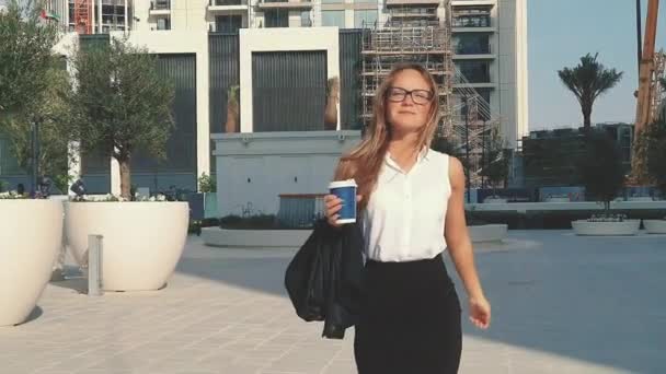 Joven mujer de negocios sonriente en vasos y taza de café caminando por la ciudad con edificios modernos en el fondo. - Metraje, vídeo