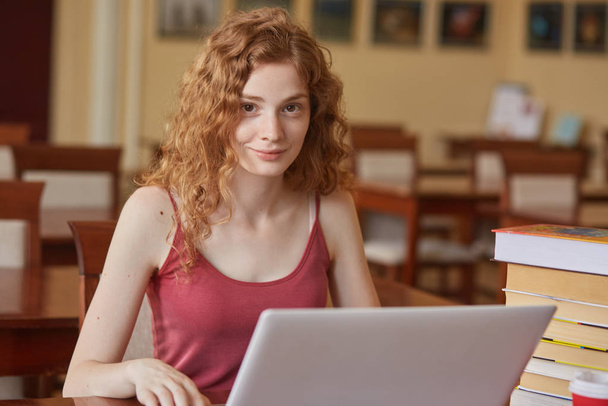 Closeup portrait of adorable sweet female having curly red hair, wearing red shirt, using laptop, sitting at library alone, smiling sincerely, being in good mood. People and studying concept. - Photo, Image