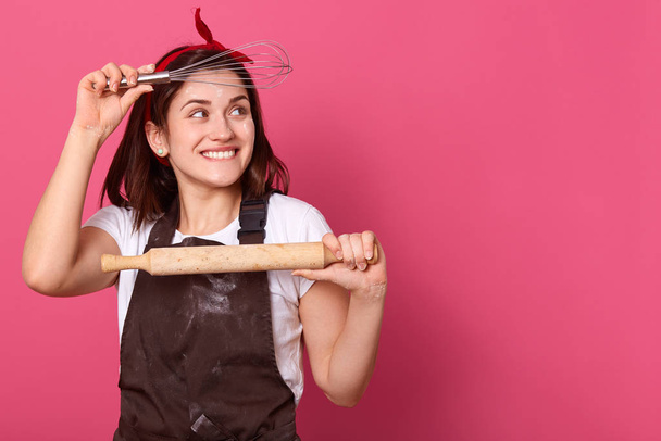 Indoor portrait of funny playful housewife looking aside, holding wooden rolling pin and whisk near face, having ideas, cooking for family, wearing apron and t shirt. Copyspace for advertisement. - Fotografie, Obrázek