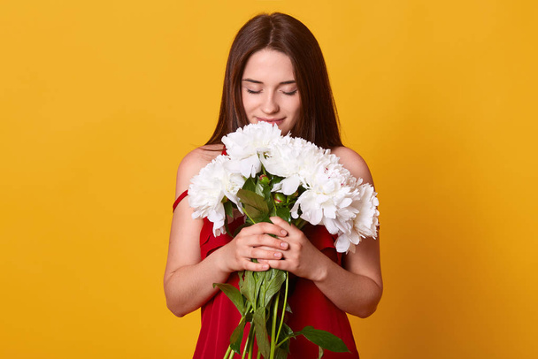 Portrait of romantic elegant brunette closing her eyes, feeling smell of flowers, holding bouquet of white peonies, standing isolated over yellow background, being delighted. Tenderness concept. - Фото, изображение