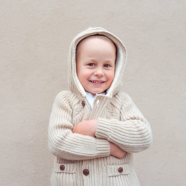 Little boy 3-5 years old, happy smiles, rejoices and laughs, background beige wall. Warm sweater with a hood. Emotions of joy of fun and relaxation at the weekend. - Fotografie, Obrázek