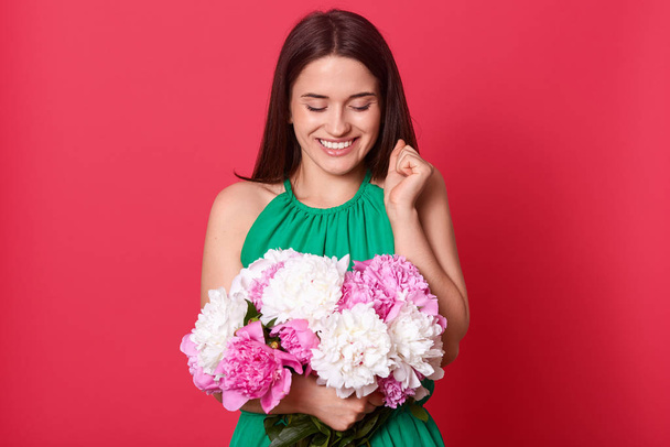 Image of cute charming adorable lady wearing green dress, raising one hand, closing eyes, celebrating day of lovers, having present, holding white and pink peonies. Saint Valentines day concept. - Valokuva, kuva