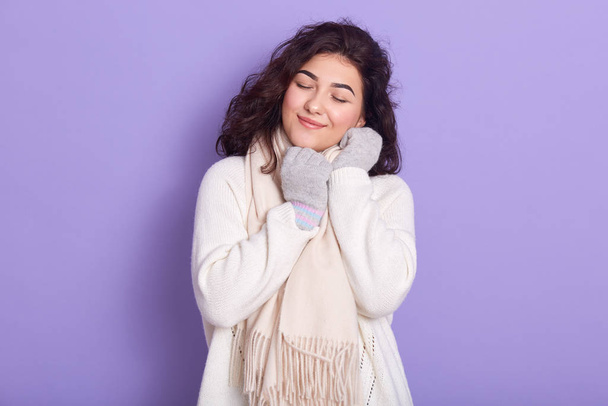 Pleasant positive beautiful female closing eyes, smiling sincerely, spending time alone, wearing white scarf, sweater and gloves, standing isolated over lilac background in studio. Winter concept. - Fotoğraf, Görsel
