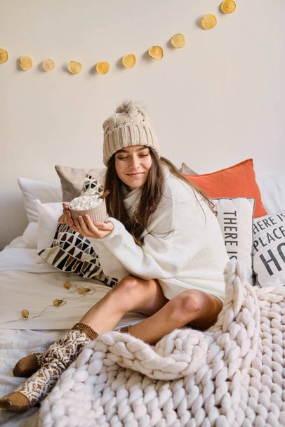 Europejska szczęśliwa dziewczyna siedzi na łóżku z filiżanką gorącego kakao z piankami. - Zdjęcie, obraz