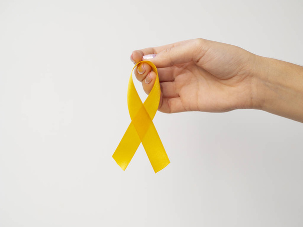 Altın kurdele beyaz arka planda parmaklarda asılı. Uluslararası Çocukluk Kanser Günü konsepti - Fotoğraf, Görsel