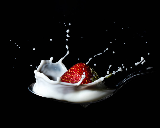 A strawberry falling down onto a spoon and splash into milk or cream - black background - Фото, изображение