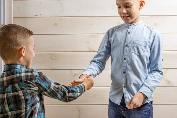 A 4-year-old boy in a blue klepy shirt cries on a light wooden background and his 10-year-old brother stands and plays rock-paper-scissors. - Fotoğraf, Görsel
