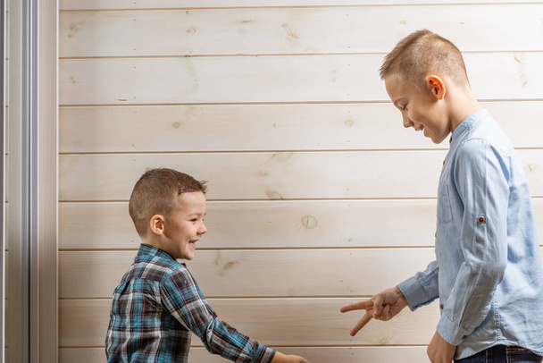 Четырехлетний мальчик в синей клеевой рубашке плачет на светлом деревянном фоне, а его 10-летний брат стоит и играет в камень-ножницы-бумагу
. - Фото, изображение