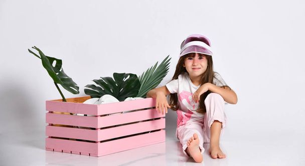 Schattig meisje naast roze houten lade zittend op de vloer in een wit t-shirt met een Flamingo in de Studio in verschillende poses - Foto, afbeelding