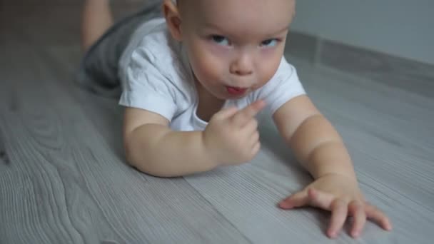 Two-year-old baby lies on the floor on his stomach and looks at the camera. Close portrait, happy childhood, blue-eyed child - Záběry, video