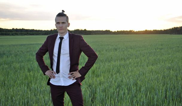 On a wheat field, a young businessman is posing for the camera with his hands on his belt in a maroon suit. Photo shoot of a young guy. - Foto, Imagem