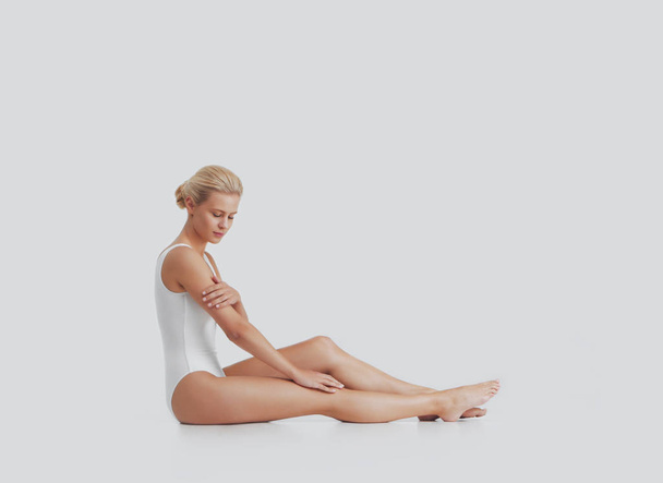 Young, beautiful, fit and natural blond woman in white swimsuit applying moisturizing cream. Massage, skin care, cellulite removal, sport and weight loss concept. - 写真・画像