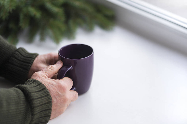 las manos de los ancianos están sosteniendo una taza de café junto a la ventana, ramas de un abeto verde sin juguetes de Navidad, vista horizontal, espacio de copia a la derecha
 - Foto, imagen