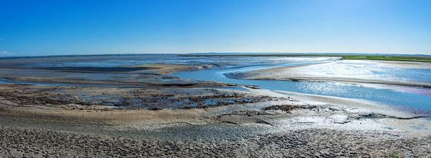 El piso de barro en la marea baja cerca de la abadía llamada Mont Saint-Michel en Normandía, Francia, la costa del Canal de la Mancha, la gente está caminando en la arena, un día soleado en verano, fondo azul cielo
 - Foto, Imagen