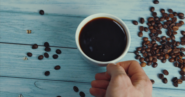 Mão masculina leva uma caneca de café. xícara de café e grãos de café. Xícara branca de café fumegante na mesa com feijão torrado
. - Filmagem, Vídeo