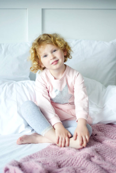 Portré kis mosolygós 5 éves lány göndör szőke haj ül rózsaszín és szürke pizsamában az ágyában. Az alvás napja. Boldog gyermekkor a kényelmes otthon a szülőkkel ellátás - Fotó, kép