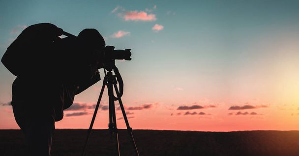 Üç ayaklı ve kameralı fotoğrafçı gün batımında geniş bir alanda manzara çeker. - Fotoğraf, Görsel