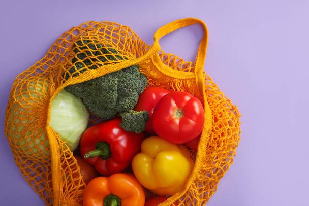 Net bag with vegetables on lilac background, top view - Foto, afbeelding