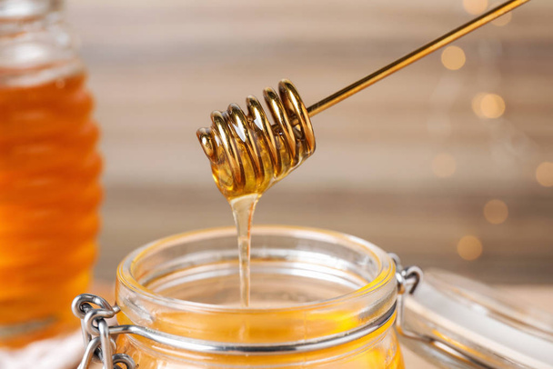 Dripping tasty honey from dipper into jar on blurred background, closeup - Fotó, kép