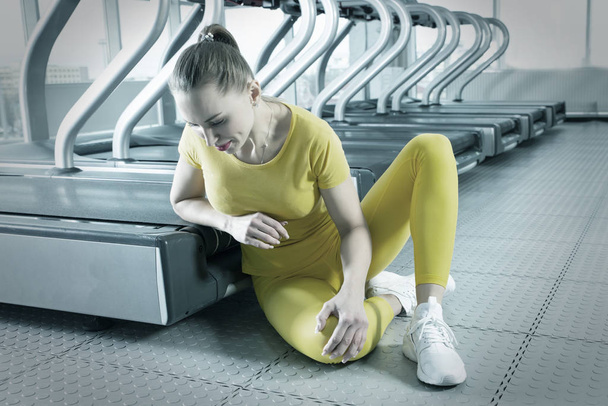 Mujer joven con la rodilla lesionada sentada en el gimnasio sintiendo dolor
 - Foto, imagen