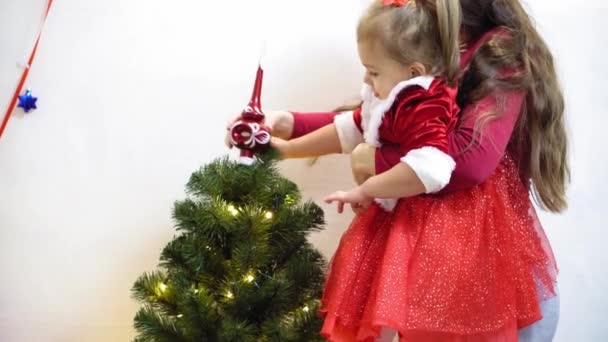 baby and pregnan mom hang red ball toy on Christmas tree. happy childhood concept. child and mother decorate tree with christmas balls. small child and a parent are playing by Christmas tree. - Metraje, vídeo