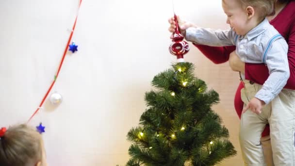 baby and pregnan mom hang red ball toy on Christmas tree. happy childhood concept. child and mother decorate tree with christmas balls. small child and a parent are playing by Christmas tree. - Filmmaterial, Video