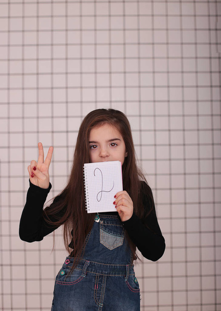 young smiling girl of eight years with long hair in a black turtleneck and denim sundress with a sign "2 two" - Photo, Image