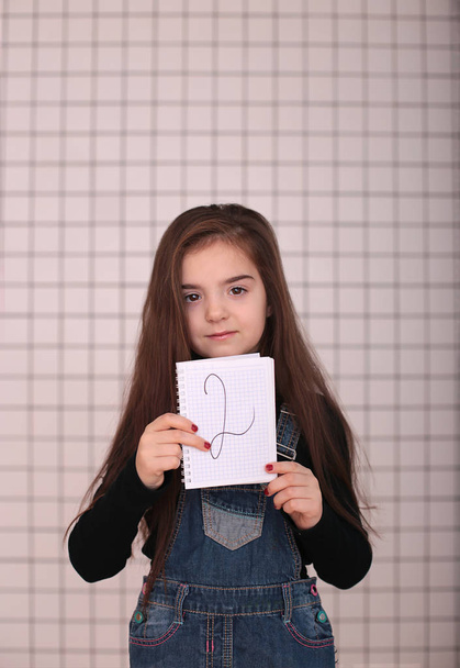 giovane ragazza sorridente di otto anni con i capelli lunghi in un dolcevita nero e denim prendisole con un segno "2 due
" - Foto, immagini