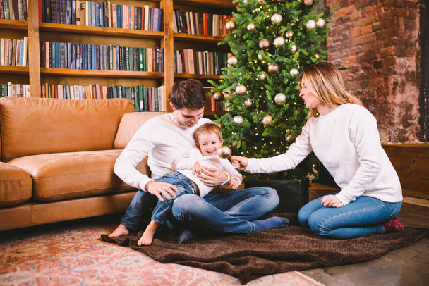 Сімейна різдвяна концепція. Прекрасна родина сидить біля ялинки в зимовий вечір. Щаслива сім'я з сином на підлозі біля дивана Вдома. Сім'я вдома в новорічний час. сім'я насолоджується новорічними святами
 - Фото, зображення