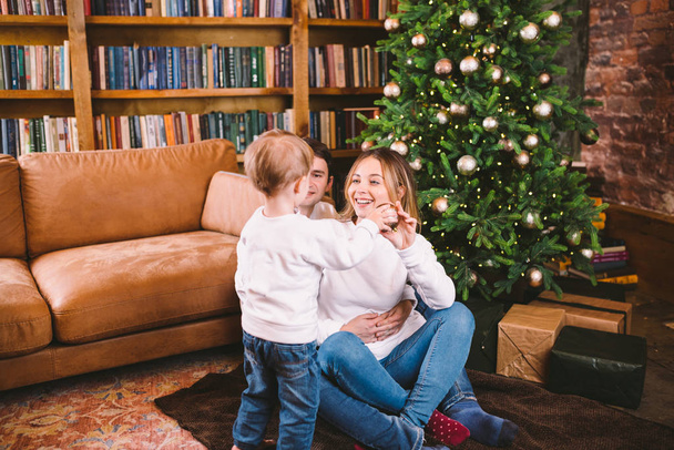 Családi karácsonyi koncepció. Szép család ül közel karácsonyfa télen este. Boldog család a fiával a padlón, közel a kanapéhoz. Család otthon, újévkor. család élvezze az újévi ünnepeket - Fotó, kép