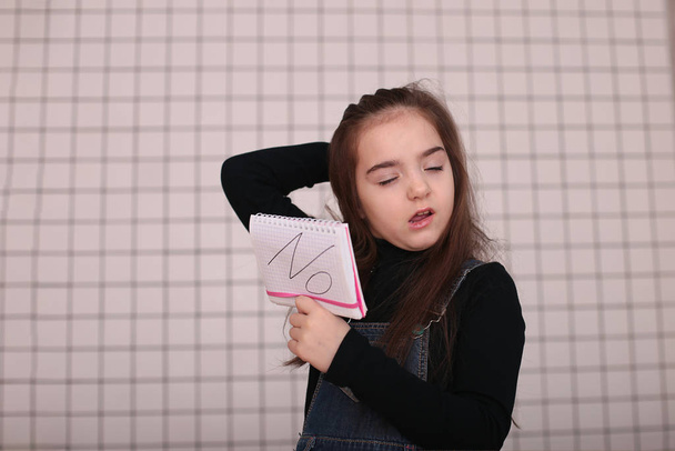 νεαρή θυμωμένη κοπέλα οκτώ ετών με μακριά μαλλιά σε μαύρο ζιβάγκο και denim sundress με την πινακίδα "No" - Φωτογραφία, εικόνα