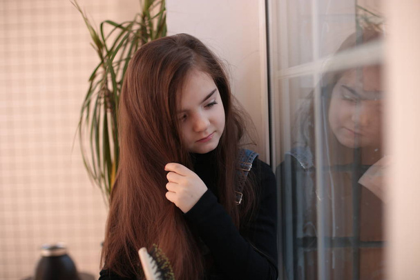 mladá usměvavá osmiletá dívka s dlouhými vlasy v černém roláku a džínsovými šaty sedí na parapetu u okna a česá si vlasy hřebenem - Fotografie, Obrázek