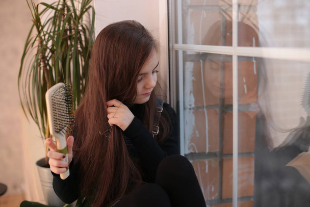 joven chica sonriente de ocho años con el pelo largo en un cuello alto negro y vaqueros sundress se sienta en un alféizar de la ventana y se peina el pelo con un peine
 - Foto, Imagen