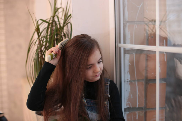 jovem sorridente menina de oito anos com cabelos longos em uma gola alta preta e jeans sundress senta-se em uma soleira da janela pela janela e penteia o cabelo com um pente
 - Foto, Imagem