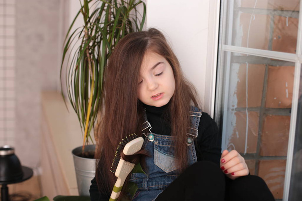 young smiling girl of eight years with long hair in a black turtleneck and jeans sundress sits on a window sill by the window and combs her hair with a comb - Foto, Imagen