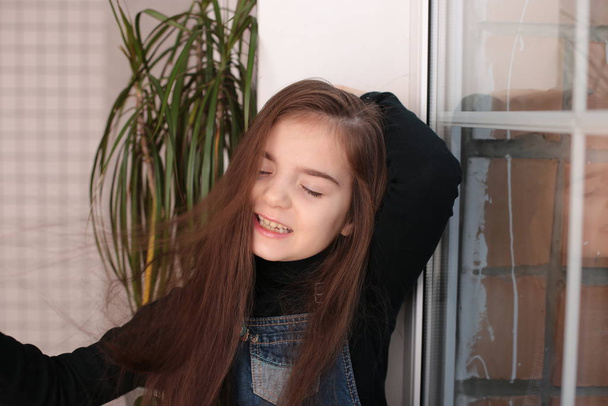 young smiling girl of eight years with long hair in a black turtleneck and jeans sundress sits on a window sill by the window and combs her hair with a comb - Zdjęcie, obraz