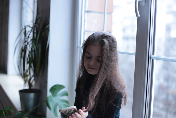 giovane ragazza sorridente di otto anni con i capelli lunghi in un dolcevita nero e jeans prendisole siede su un davanzale vicino alla finestra e pettina i capelli con un pettine
 - Foto, immagini