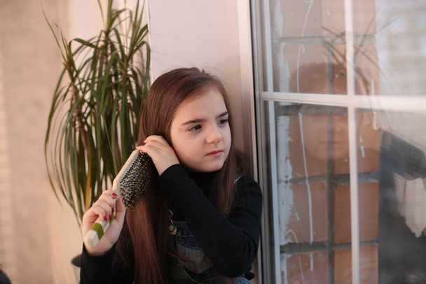 jong lachend meisje van acht jaar met lang haar in een zwarte coltrui en jeans sundress zit op een vensterbank bij het raam en kamt haar haar met een kam - Foto, afbeelding