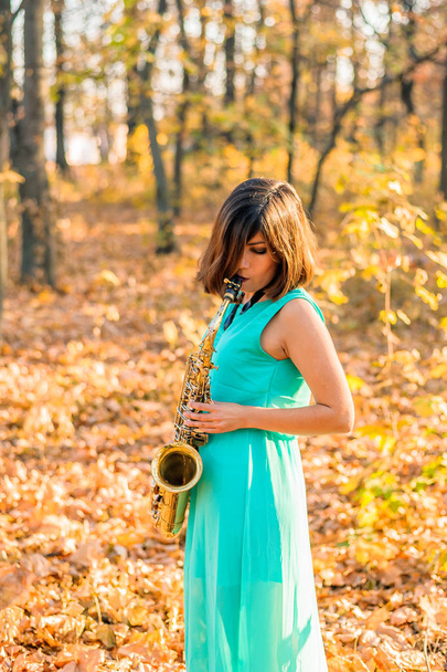 Güzel bir vücudu ve siyah saçları olan bir kız, sarı bir sonbahar parkında elinde alto saksafonuyla uzun mavi bir elbiseyle dikiliyor. - Fotoğraf, Görsel