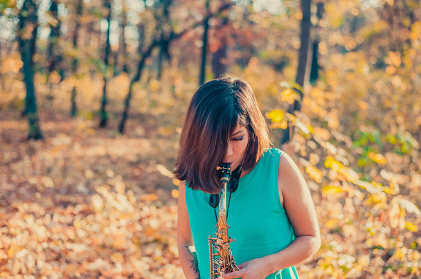 uma menina com uma bela figura e cabelo preto olha para longe, de pé em um parque de outono amarelo em um vestido azul longo com um saxofone alto em suas mãos
 - Foto, Imagem