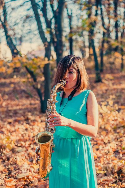 dziewczyna z piękną figurą i czarnymi włosami odwraca wzrok, stojąc w żółtym jesiennym parku w długiej niebieskiej sukience z saksofonem altowym w rękach - Zdjęcie, obraz
