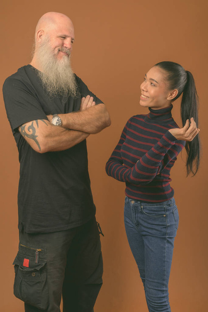 Genç Asyalı transseksüel bir kadınla kahverengi arka planı olan olgun sakallı kel bir adam. - Fotoğraf, Görsel