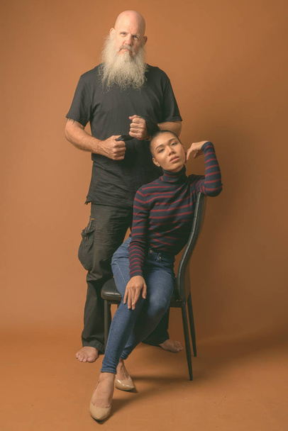 Зрелый бородатый лысый мужчина с молодой азиатской трансгендерной женщиной на коричневом фоне
 - Фото, изображение