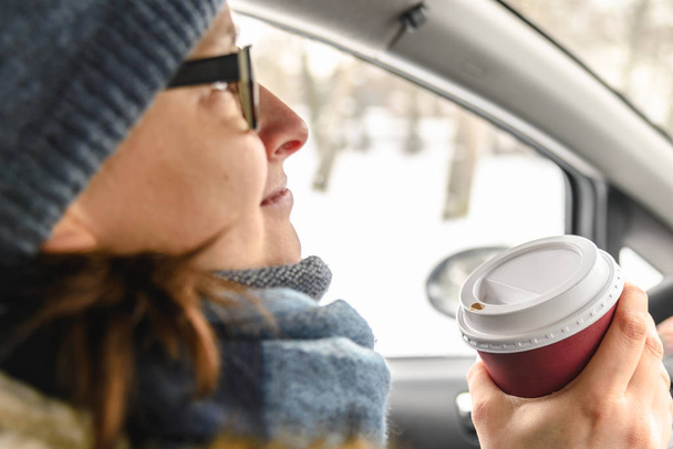 Woman driver drinking coffee in the car, driving and holding cup of hot beverage inside the auto in winter, snow behind window - Foto, afbeelding