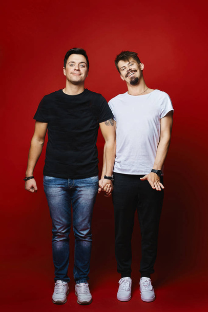 Full length portrait of two cheerful young men wich smiling and holding hands, isolated on a red background. Two handsome guys with modish hairstyles and in stylish outfits. Concept of friendship - Foto, Imagen
