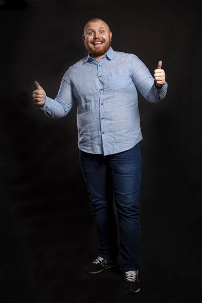 A fat red-haired man with a beard and mustache in a blue shirt and jeans stands holding thumbs up and laughs. Looks at the camera. Black background.  - Photo, image